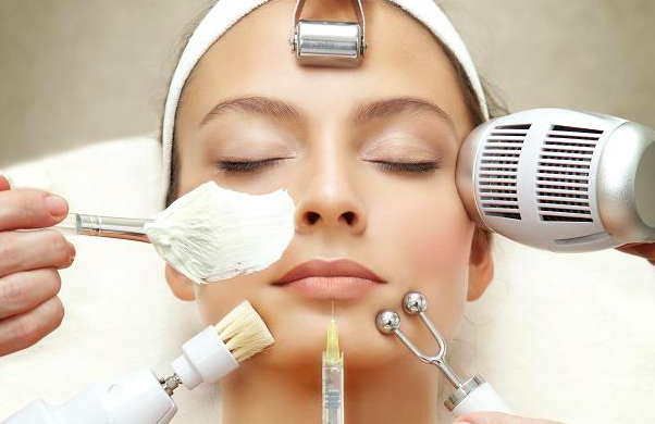 Woman receiving a multi-step facial treatment with cream, roller, brush, syringe, and electric devices near her face.