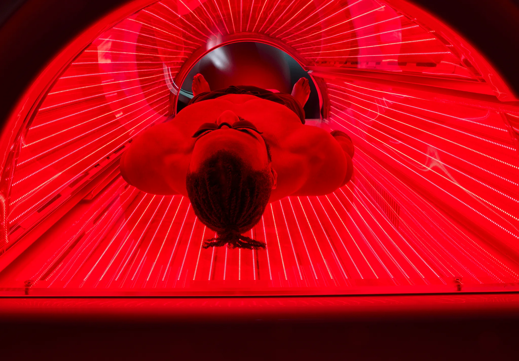 Top view of a shirtless man with braided hair inside a red light therapy bed glowing with red LED lights.