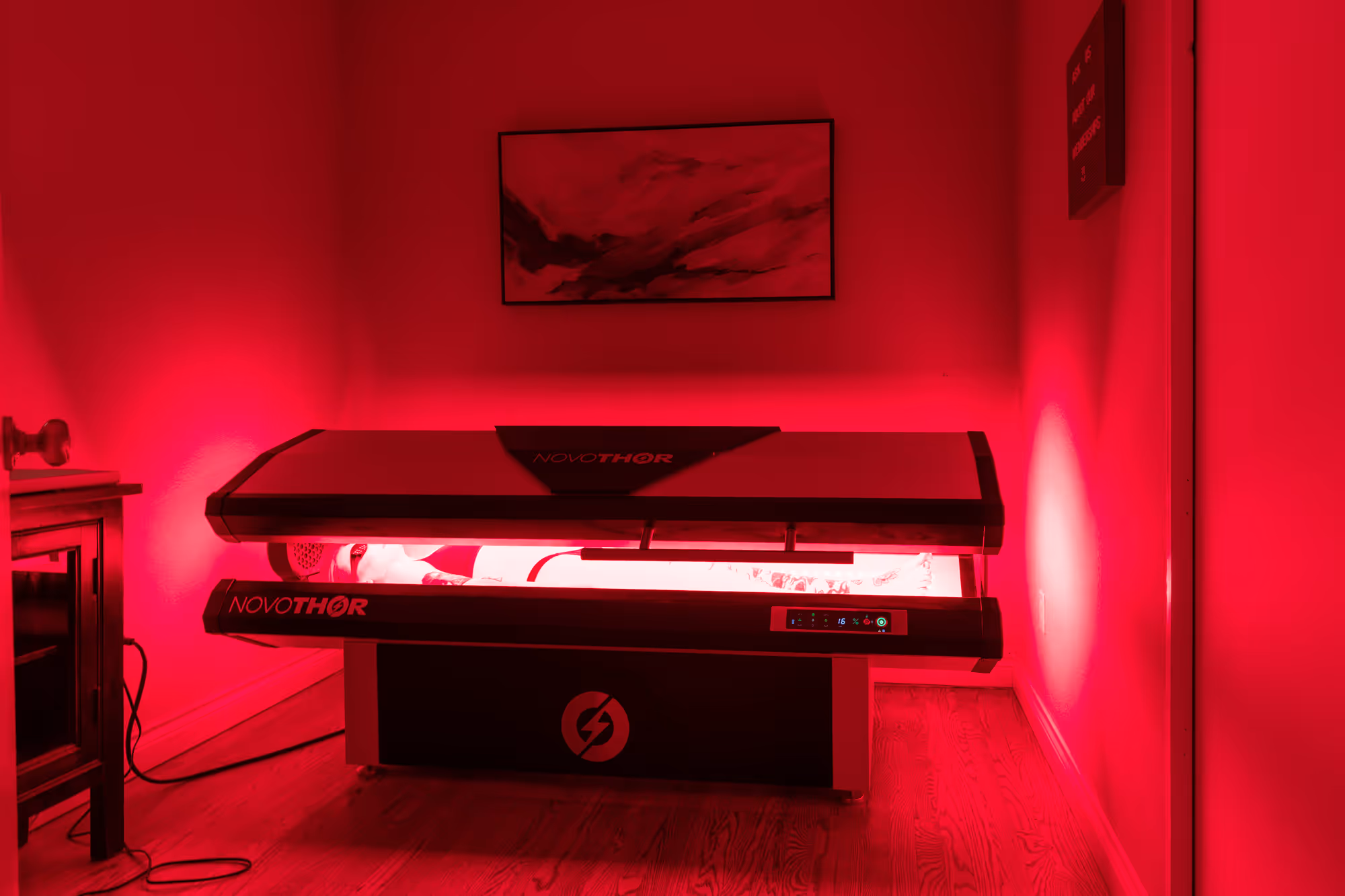 Person lying inside a NovoTHOR light therapy pod emitting red light in a wooden floor room.
