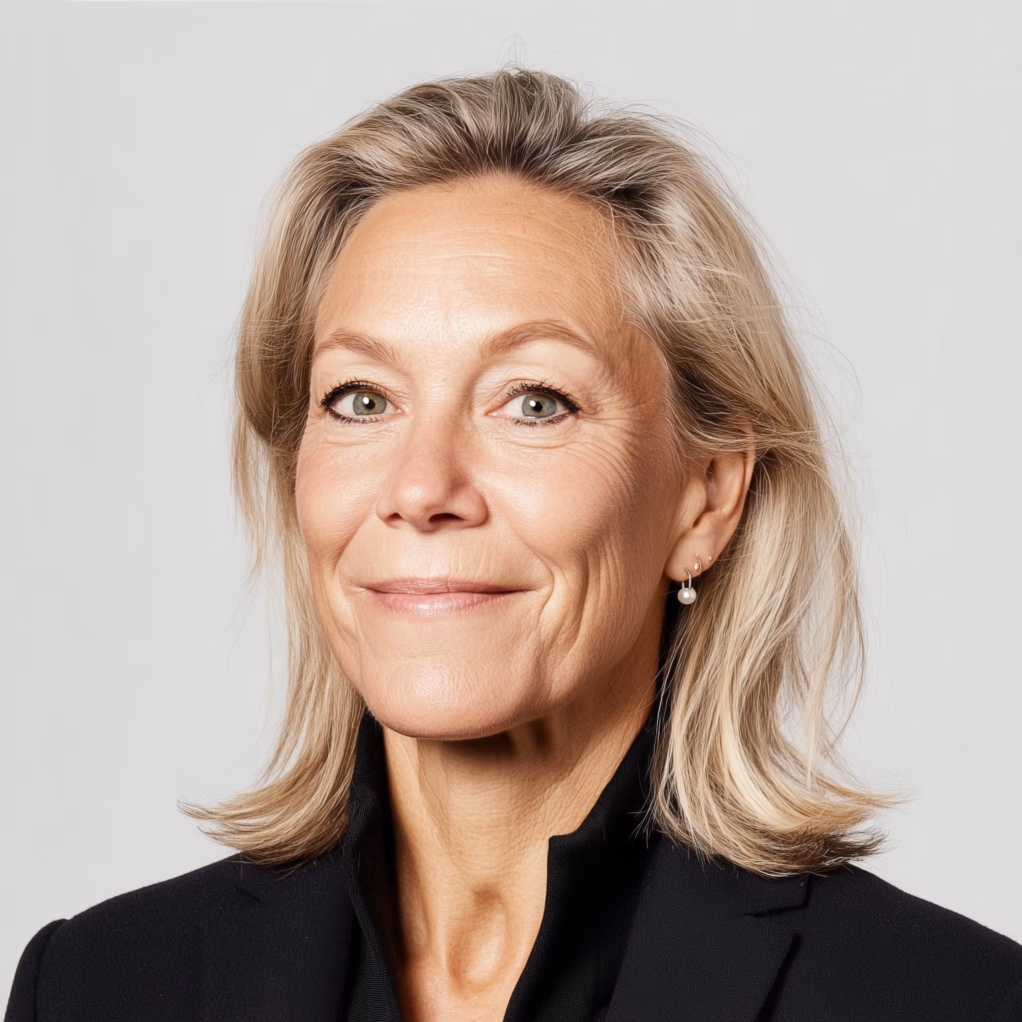 Portrait of a confident middle-aged woman with light blonde hair, green eyes, and pearl earrings wearing a black blazer.