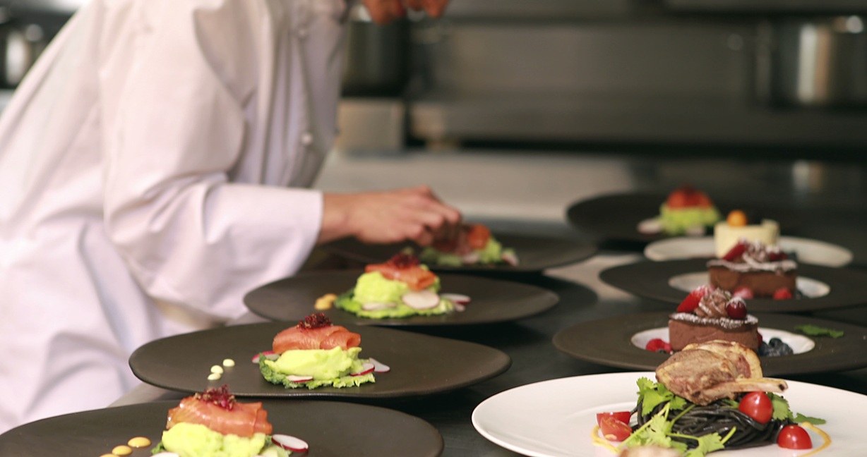 Chef in white uniform plating gourmet dishes with salmon, avocado puree, and desserts on black and white plates.