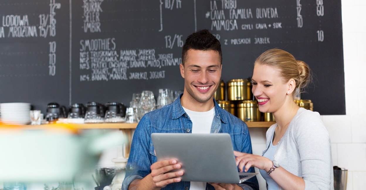 Smiling man and woman looking at a laptop together inside a cafe with a menu board in the background.