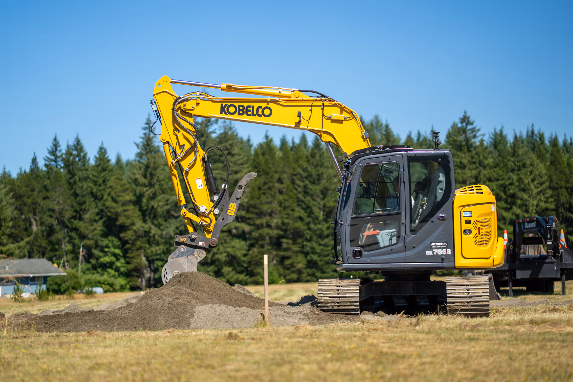 Yellow Kobelco excavator digging soil on a grassy field with trees in the background under a clear blue sky.