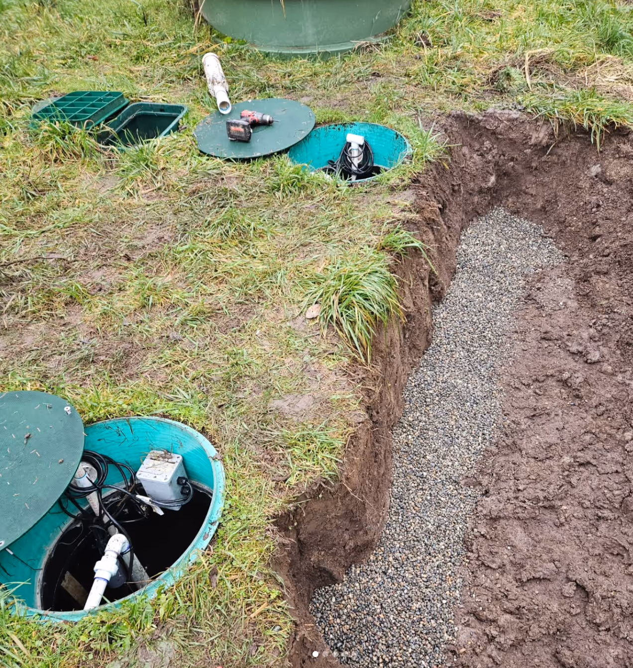 Open green septic tanks with tools nearby and a trench filled with gravel next to them in a grassy area.