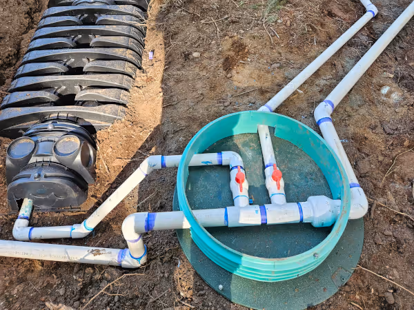 PVC piping system with two red-handled valves inside a green circular valve box on dirt ground.