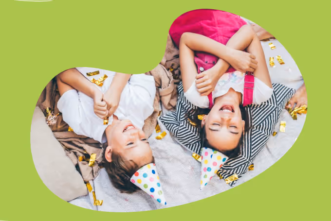 Two children lying on a blanket wearing party hats, laughing and holding hands with golden confetti scattered around.