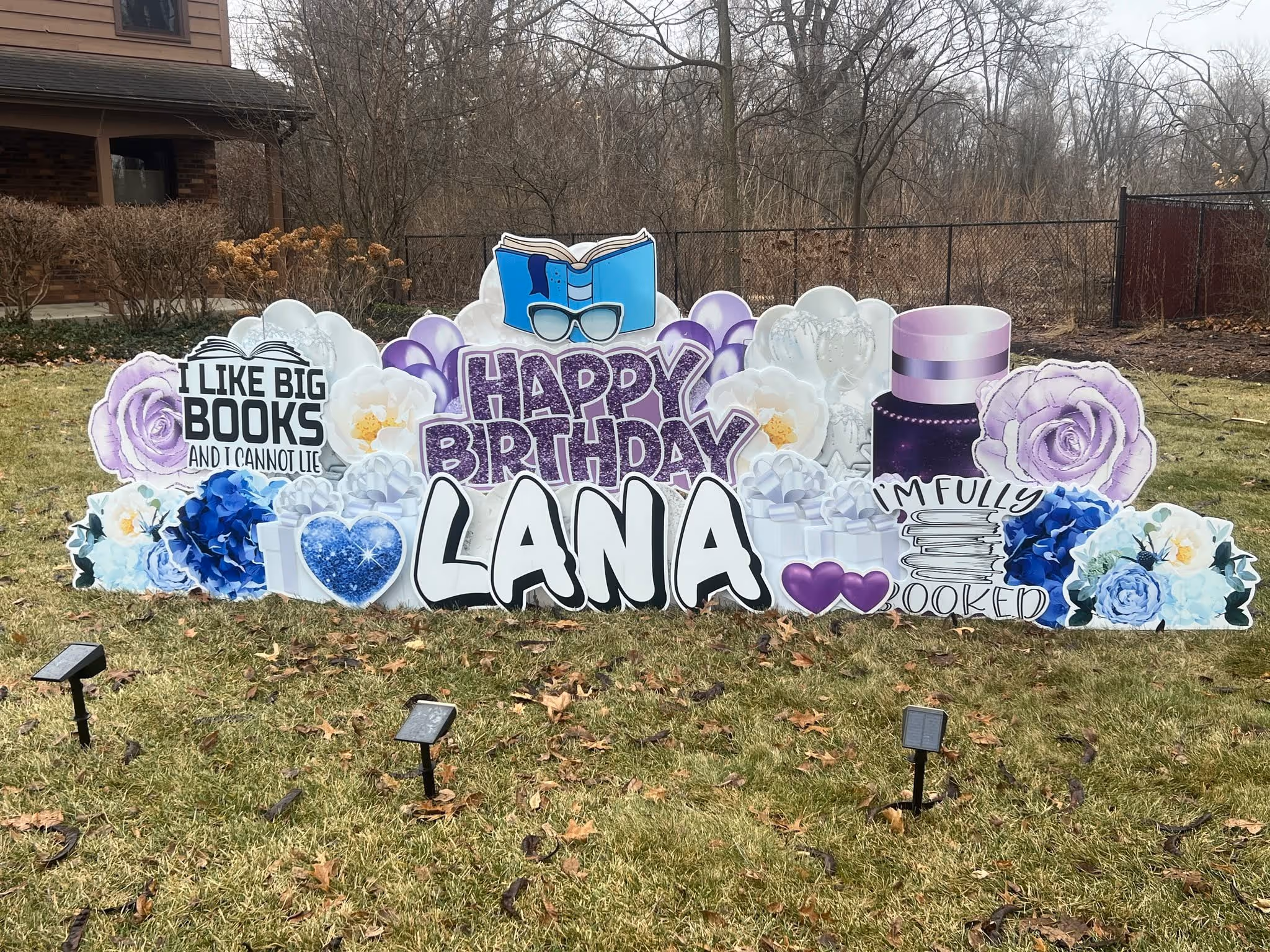 Outdoor birthday yard sign with purple and white flowers, books, and text saying Happy Birthday Lana, I like big books and I cannot lie, and I'm fully booked.
