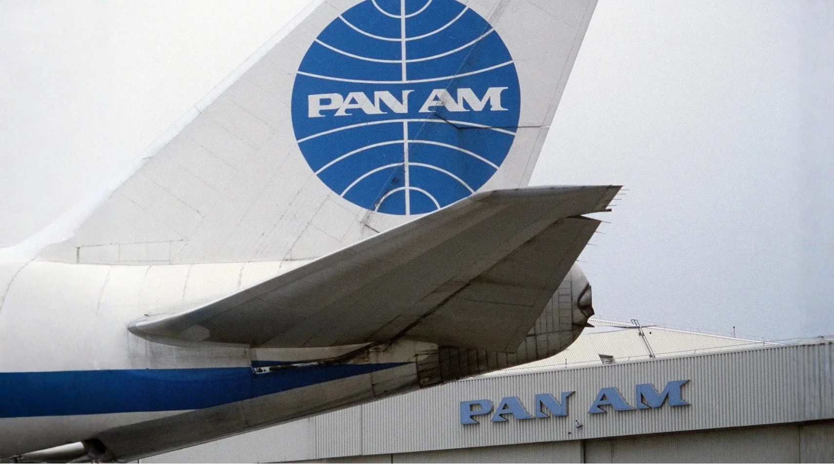Tail section of a Pan Am airplane with the Pan Am logo and a building in the background also displaying the Pan Am name.