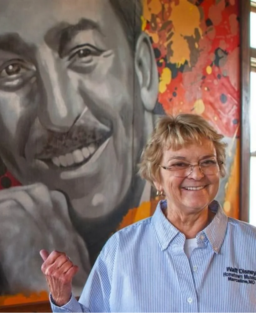 Smiling older woman wearing glasses and a striped blue shirt standing in front of a large black-and-white mural of a man with a mustache.