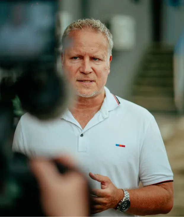 Middle-aged man with short blond hair in a white polo shirt speaking in front of a camera.