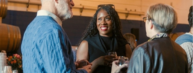 Three people holding drinks and smiling while engaging in conversation at an indoor event.