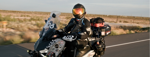 Person in full motorcycle gear riding a touring motorcycle on a road with a desert landscape in the background.