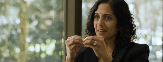 Woman with curly dark hair speaking and gesturing with her hands by a window with blurred greenery outside.