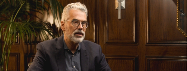 Older man with white hair and beard wearing glasses and a dark blazer sitting in a wood-paneled room with green plants.
