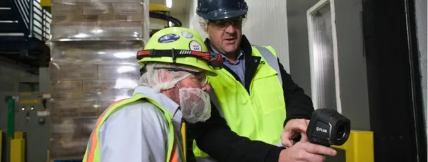 Two workers wearing safety gear inspecting a thermal camera device inside a warehouse.