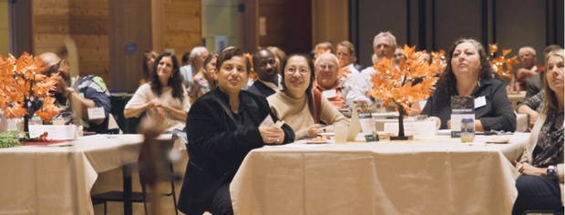 Group of diverse people sitting at banquet tables decorated with autumn-themed centerpieces during an indoor event.