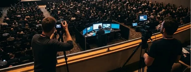 Two camera operators filming a large audience seated in an auditorium with a person managing multiple monitors at a control desk.