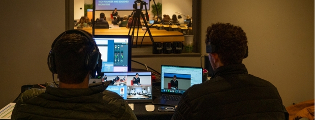 Two people wearing headphones operating multiple laptops and video equipment to live stream an event in a dimly lit room.