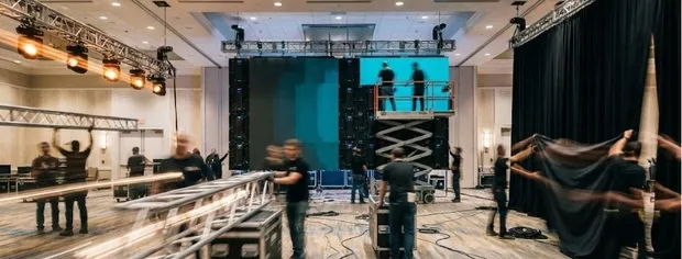 Blurry team setting up lighting, trusses, and black curtains in a large indoor event space.
