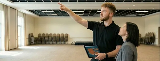 A man holding a tablet points while a woman attentively looks in the same direction inside a large empty room with stacked chairs.
