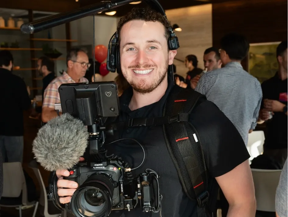 Smiling cameraman holding a professional camera rig with microphone and monitor in a crowded indoor setting.