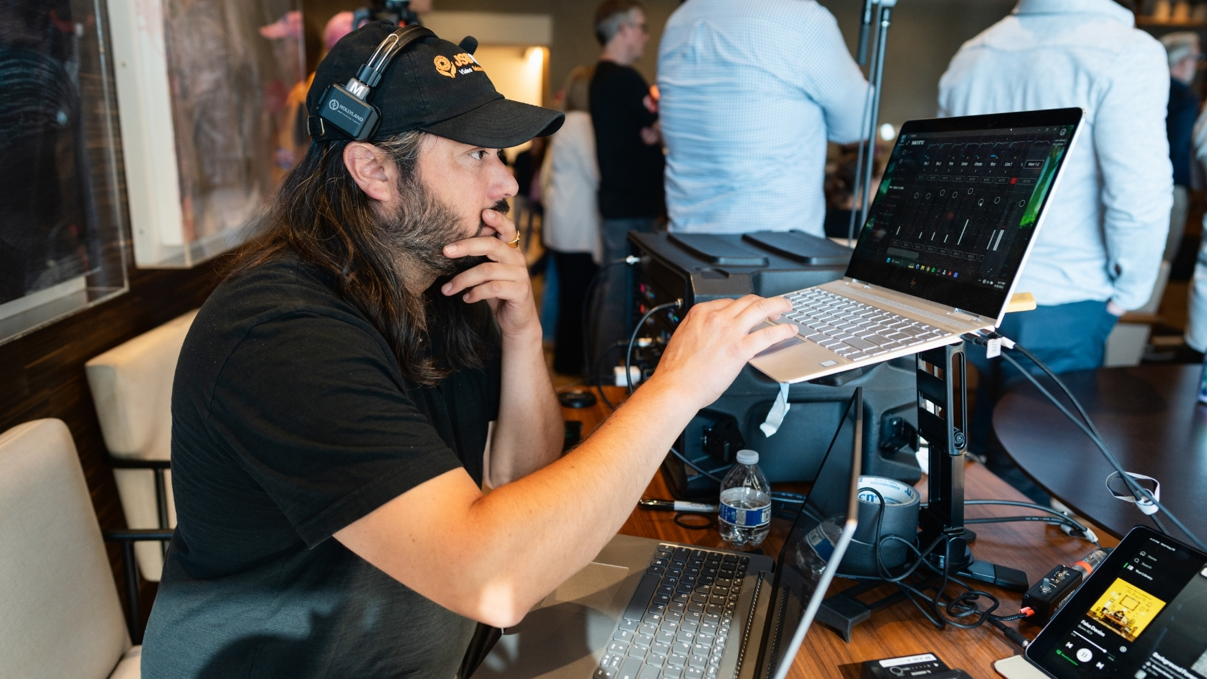 Man with long hair and cap wearing headphones, working on two laptops side by side in a busy room.