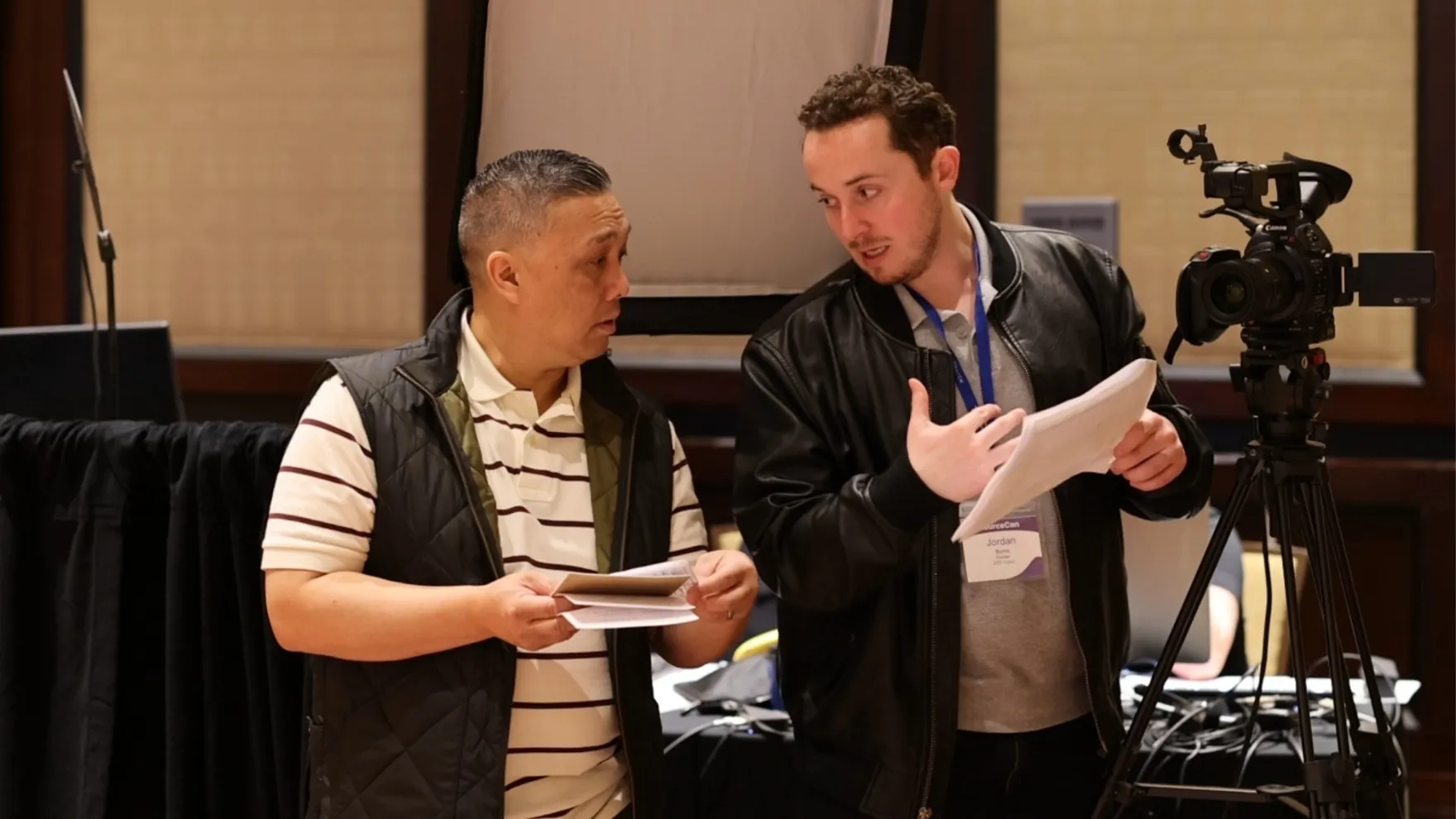 Two men discussing notes beside a professional video camera on a tripod indoors.