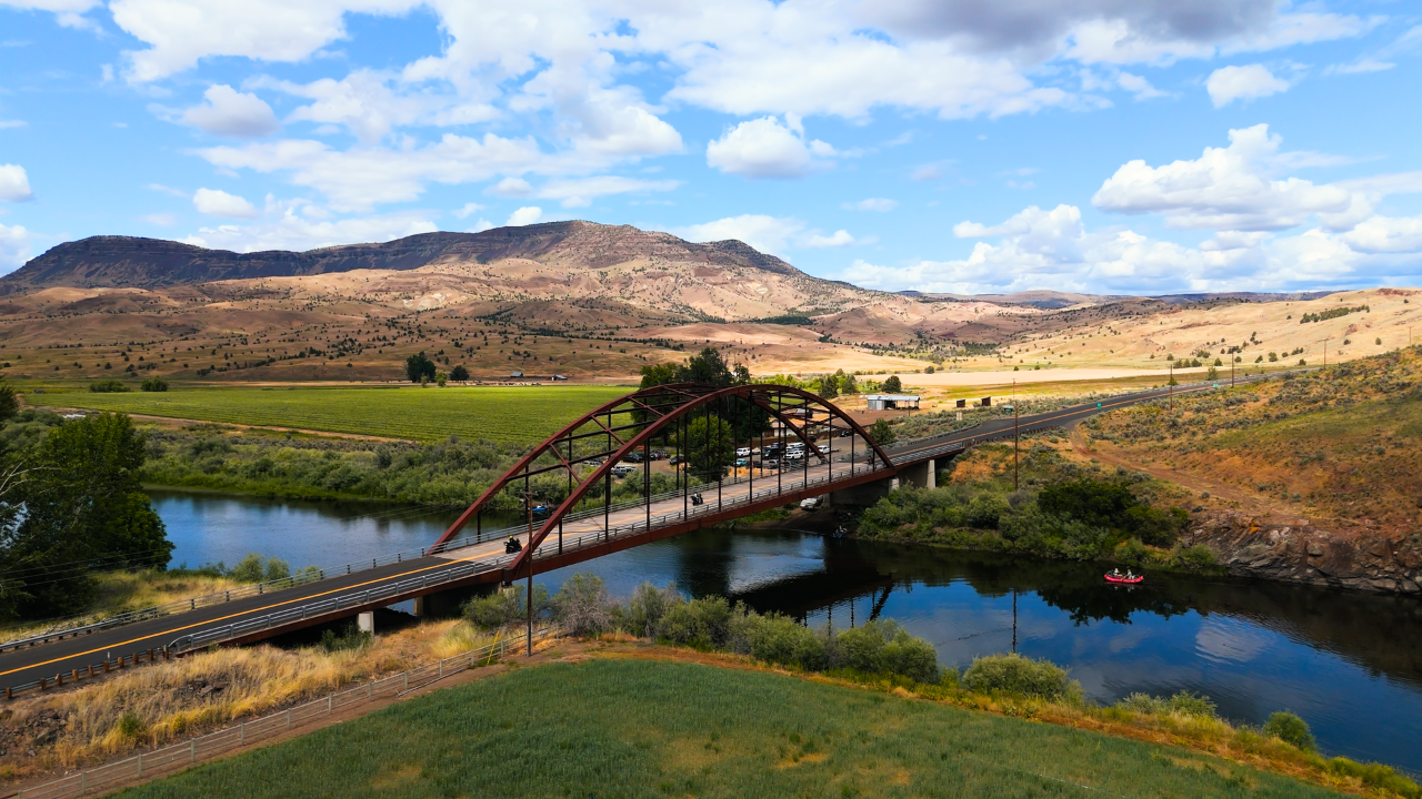 Arched metal bridge over a calm river with motorcyclists crossing, surrounded by fields and rolling hills under a partly cloudy sky.