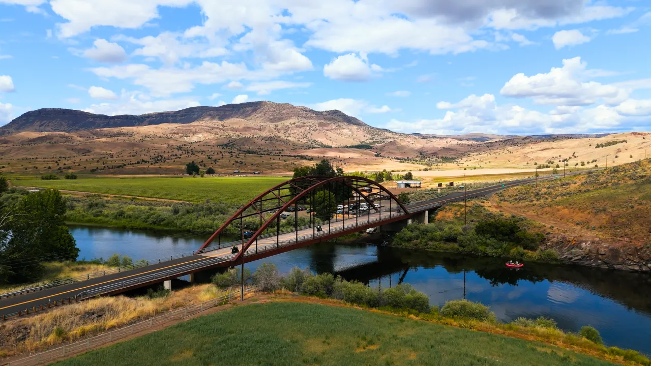 Arched metal bridge over a calm river with motorcyclists crossing, surrounded by fields and rolling hills under a partly cloudy sky.