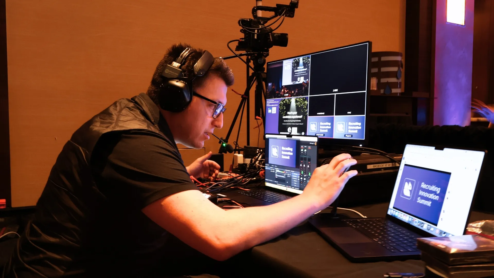 Man wearing headphones and glasses operating multiple laptops and a monitor for a live event streaming setup.