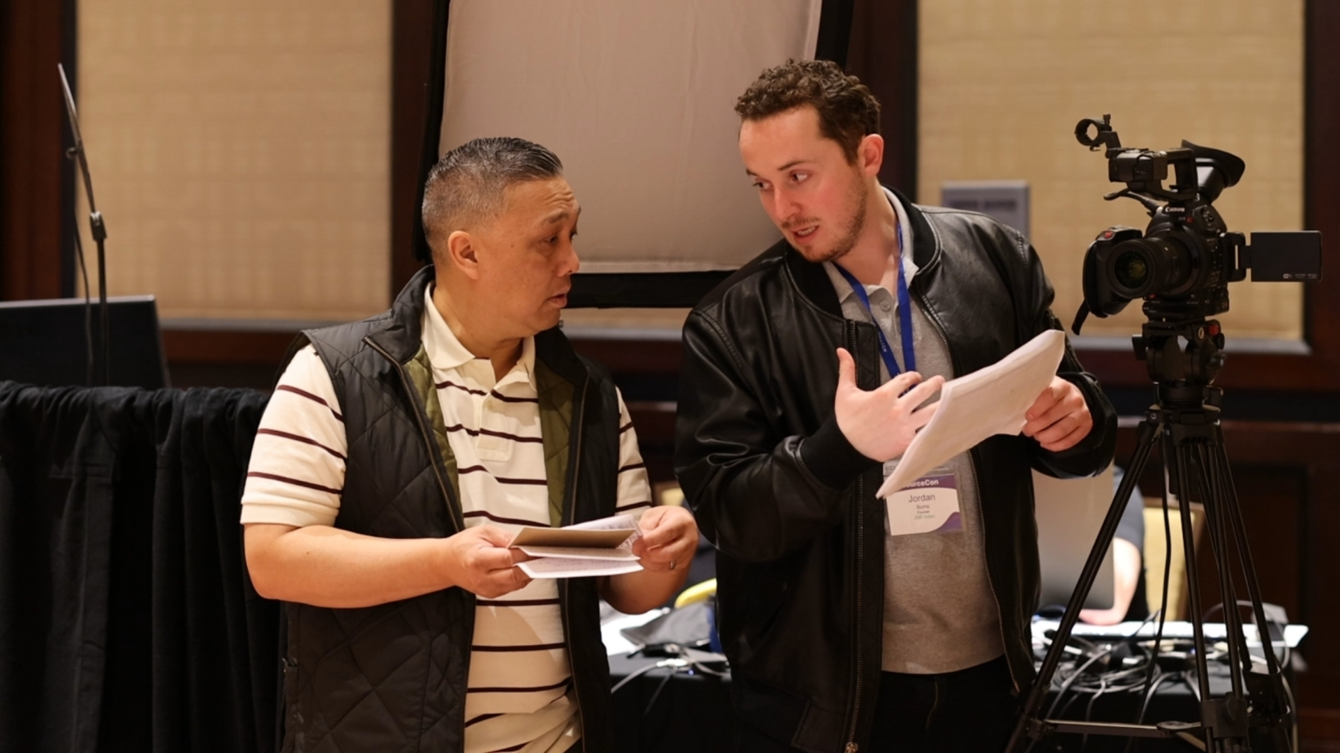 Two men discussing with papers in front of a video camera on a tripod during a recording or interview setup.