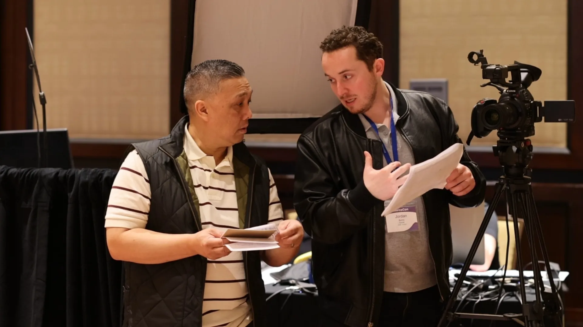 Two men discussing with papers in front of a video camera on a tripod during a recording or interview setup.