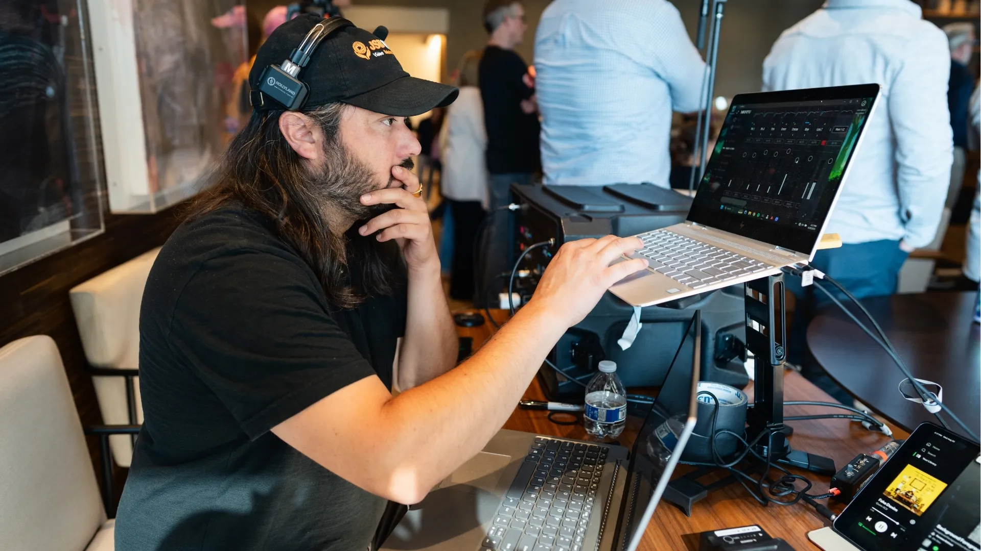 Man wearing a black cap and headphones operates a laptop on a stand at a crowded event.