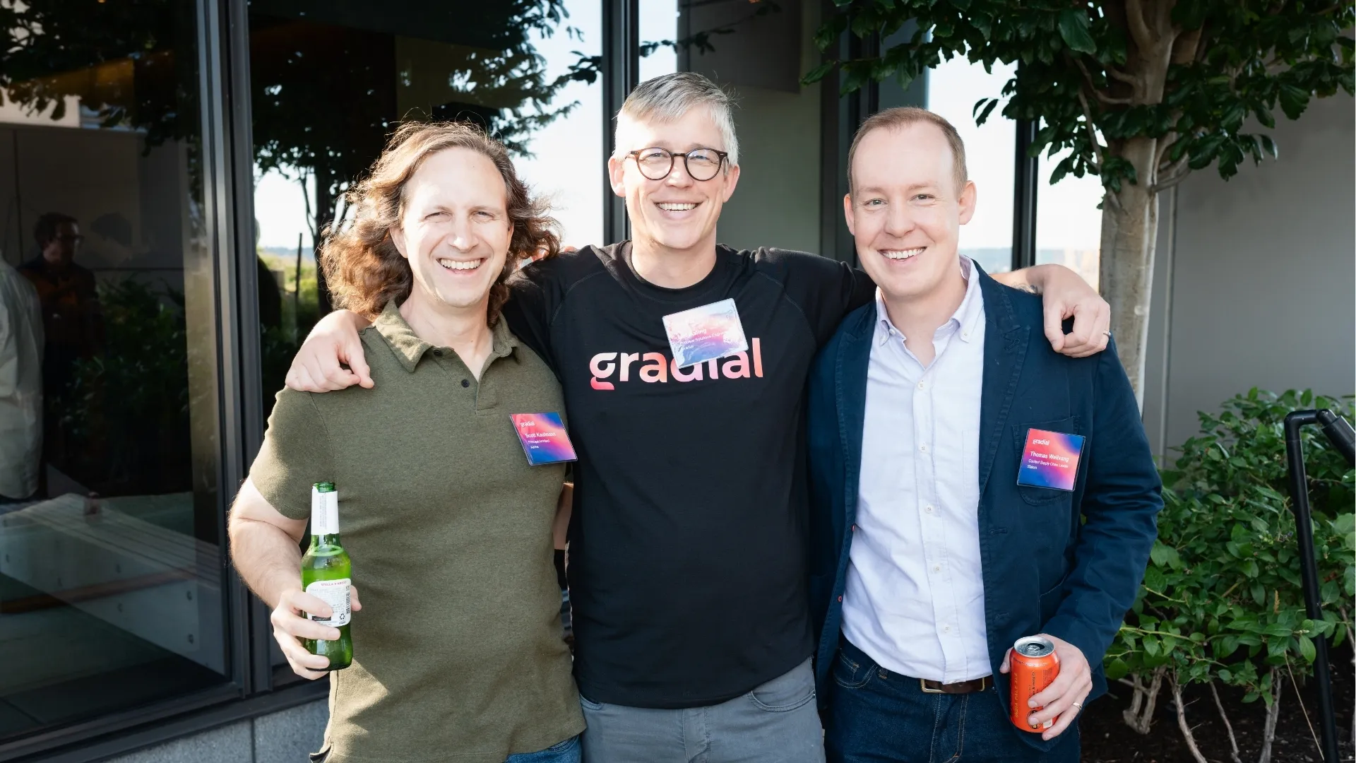 Three men smiling with arms around each other at an outdoor event, each holding a drink and wearing event badges.