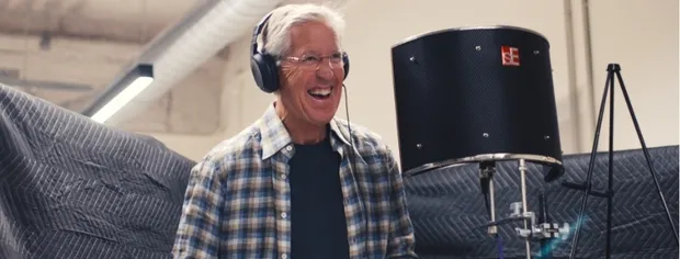 Older man wearing headphones and glasses smiling in front of a microphone in a recording studio.