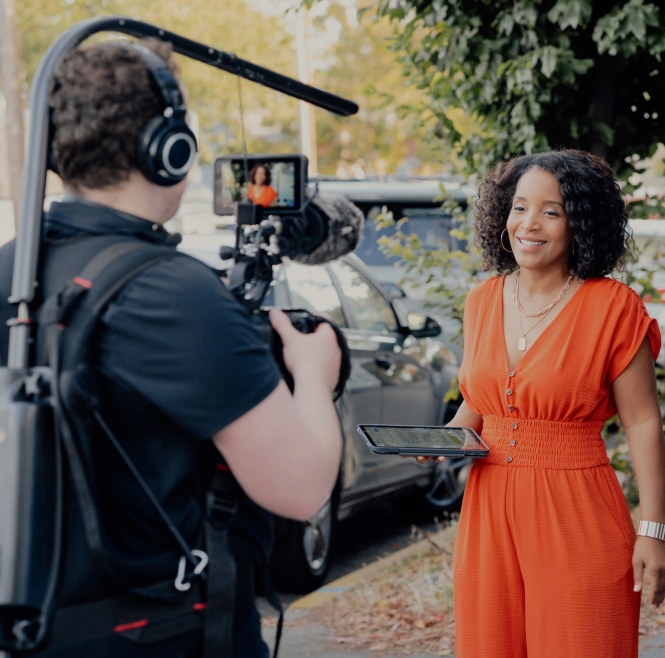 Woman in orange dress holding a tablet being filmed by a cameraman with headphones and microphone.