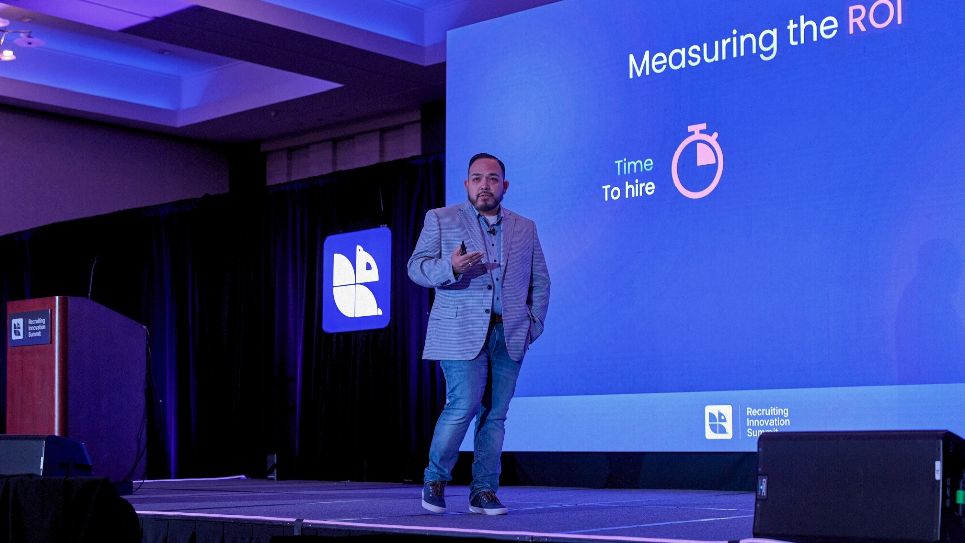Conference presenter onstage discussing time-to-hire and ROI metrics during a Recruiting Innovation Summit keynote