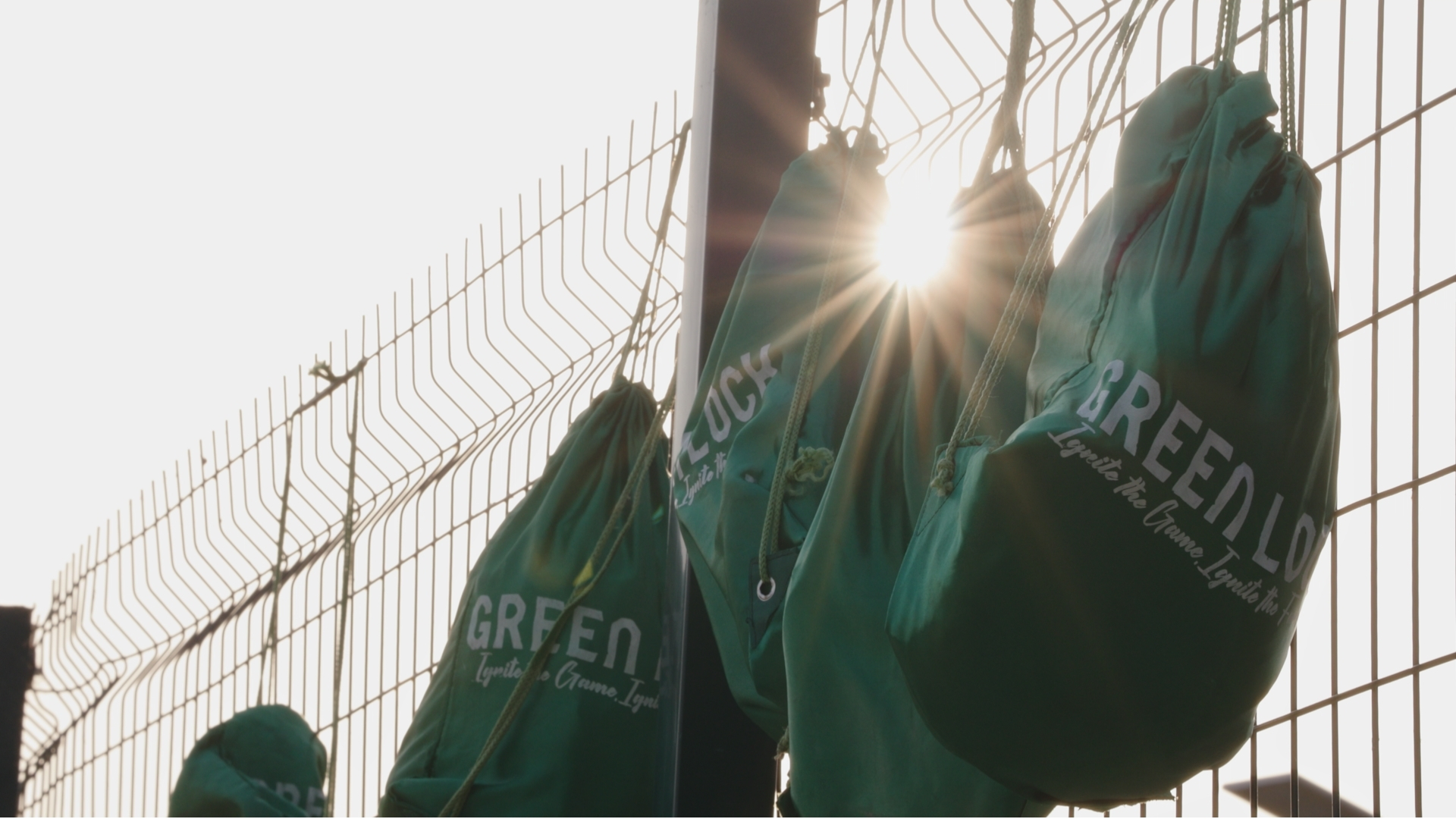 Green Lock FC kit bags hanging on a metal fence as sunlight filters through during an outdoor training session