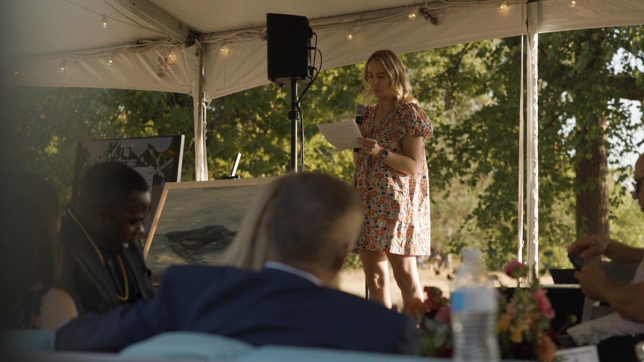 Woman speaking at outdoor tent event with audience and artwork displayed