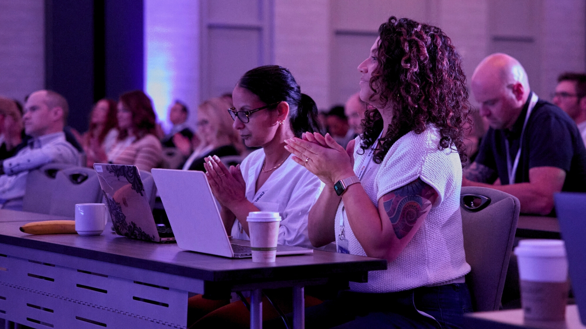 Women with laptops at a conference table under purple lighting, applauding speaker