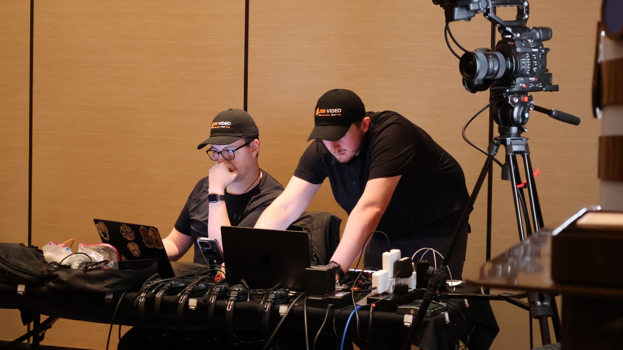 Two video professionals wearing branded caps monitor equipment during a film production setup.