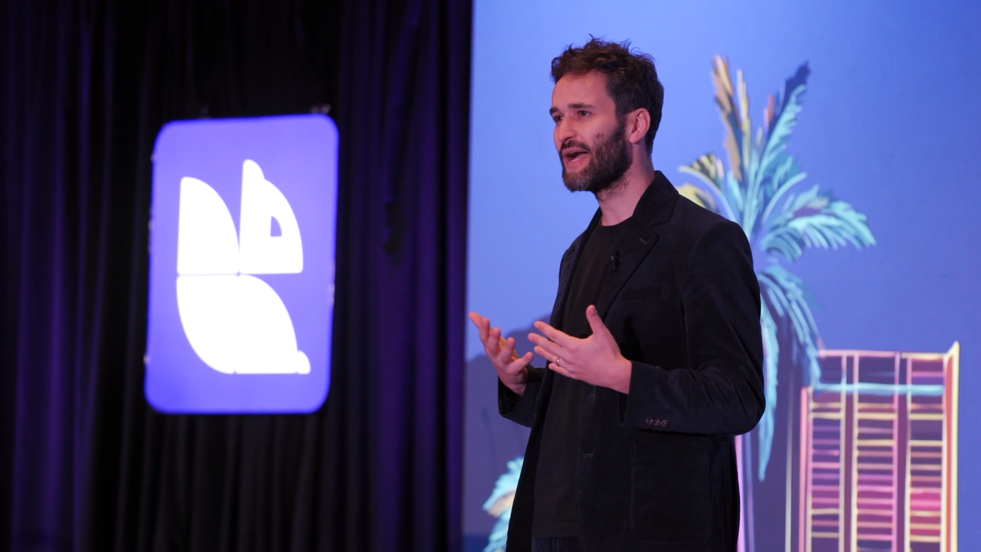 Man in black blazer speaking on stage with logo display and plant decor