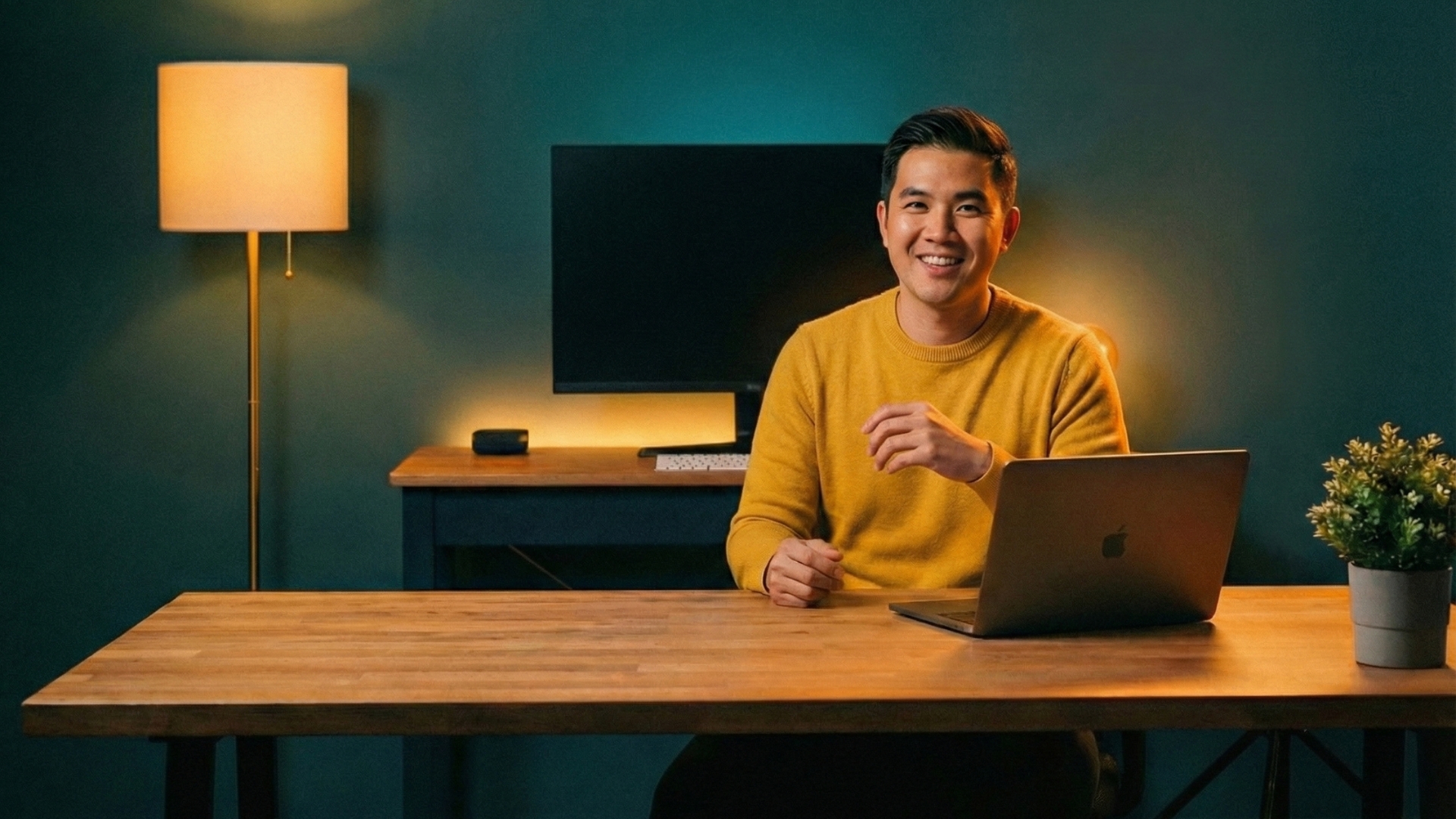 Smiling man in yellow sweater at wooden desk working on laptop with warm lighting setup