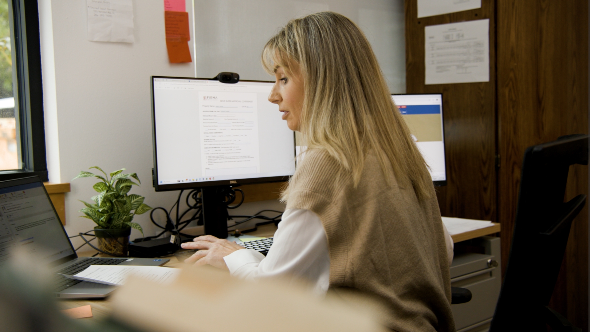 Woman working at desk with multiple monitors and plant in office