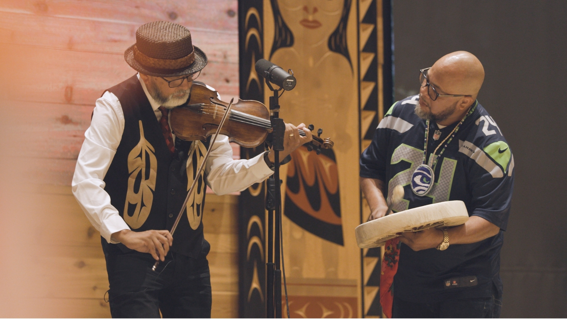 Two musicians perform on stage: elder man playing violin in traditional vest, younger man with drum
