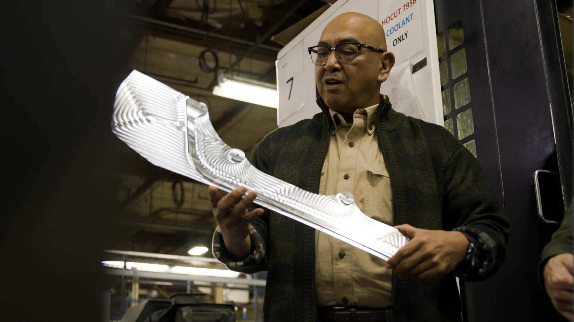 Man in glasses and cardigan holds and examines a white textured component in industrial workshop setting.