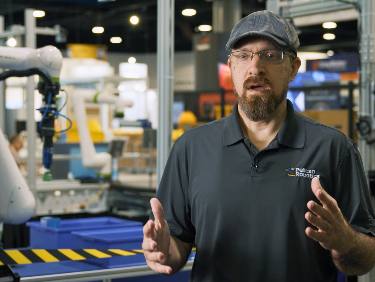 Presenter speaking on camera from a robotics exhibit at a trade show.