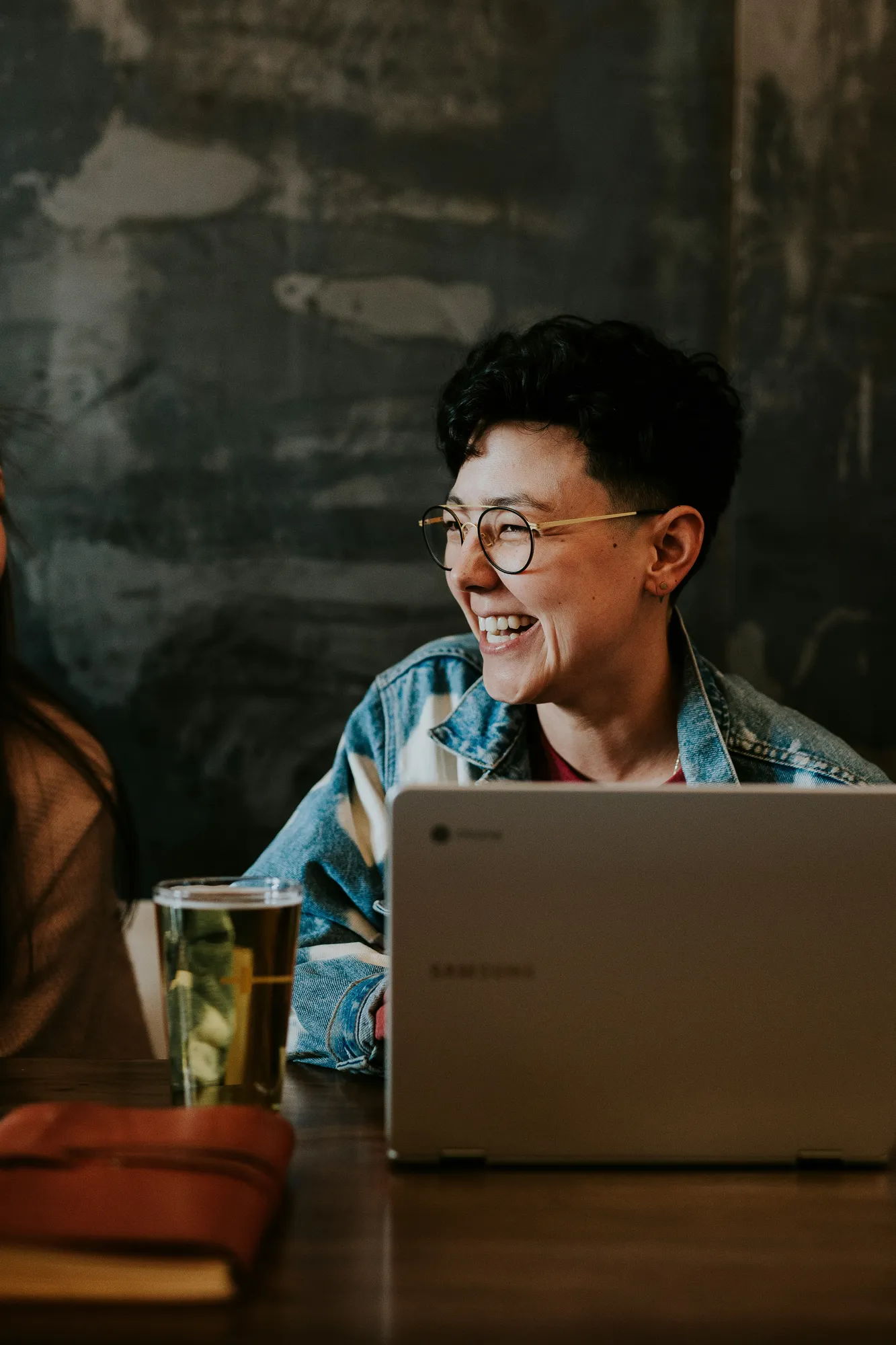 Person with short curly hair and round glasses smiling while sitting at a table with a laptop and a glass of beverage.