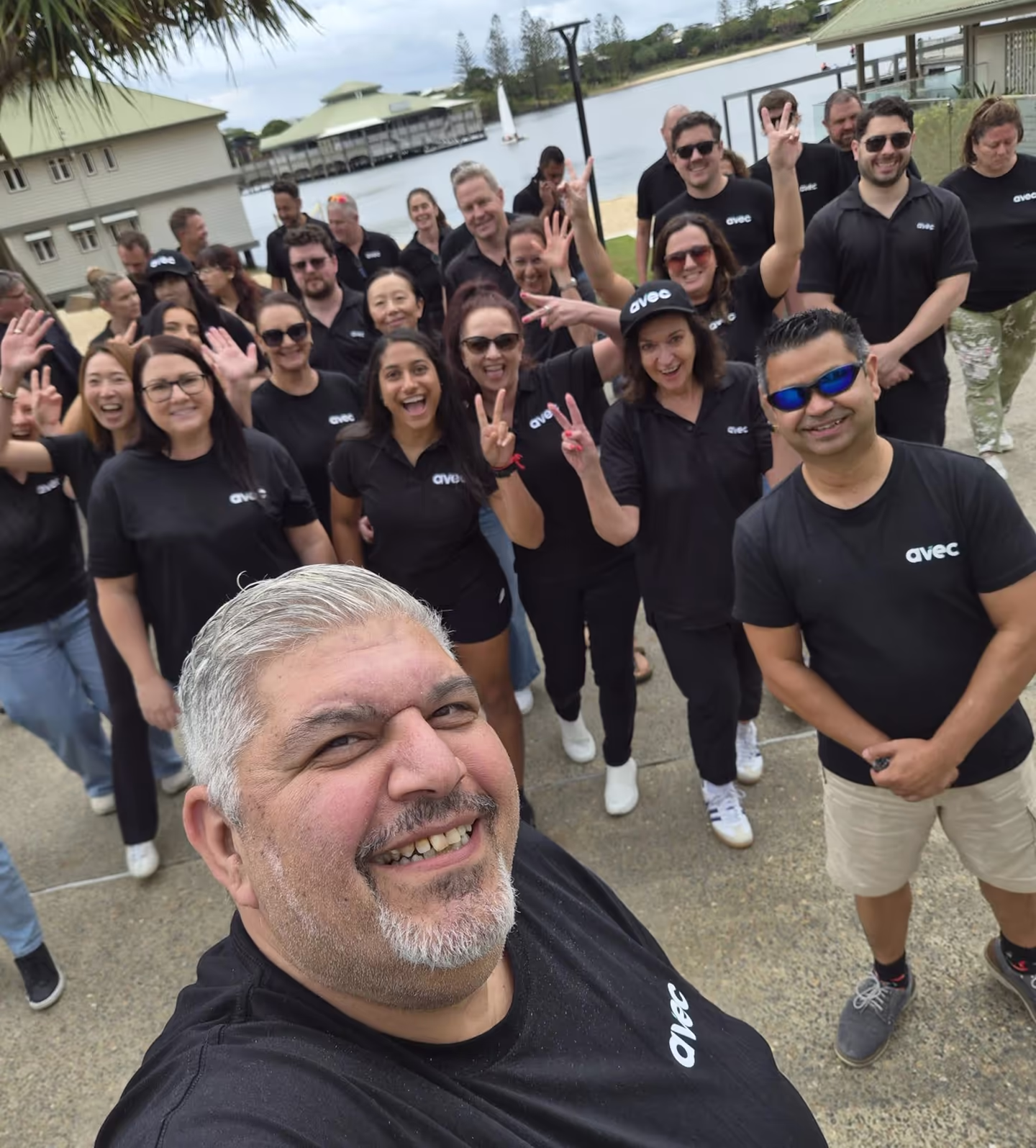 Group of smiling people wearing black Avec shirts posing for a selfie outdoors near water.