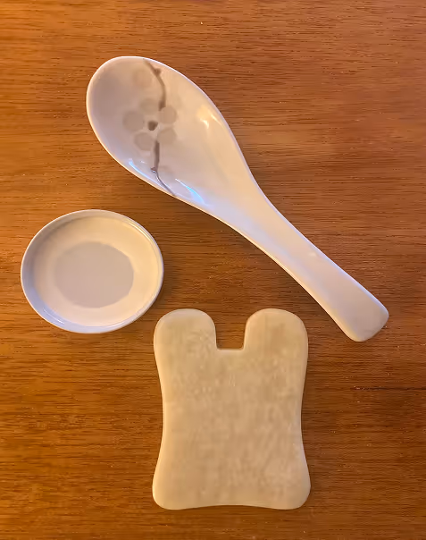 A ceramic spoon with floral design, a round saucer, and a jade gua sha tool are arranged on a wooden surface, conveying a serene and elegant tone.